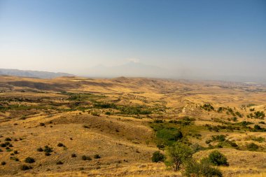 Uçsuz bucaksız, kuru ve çorak bir çöl manzarası. Birkaç ağaç etrafa saçılmış. Gökyüzü açık ve mavidir ve güneş ışıl ışıl parlar.