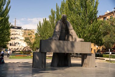 Arkasında bir bankla masada oturan bir adam heykeli. Heykel ağaçlar ve bir parkla çevrili.