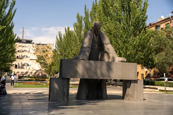 Arkasında bir bankla masada oturan bir adam heykeli. Heykel ağaçlar ve bir parkla çevrili.