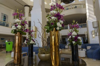 Merkezinde büyük bir çiçek aranjmanı olan bir otel lobisi. Çiçekler mor ve beyazdır ve farklı boyutlarda üç vazo vardır. Vazolar masaya konur.