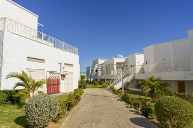 Kırmızı kapısı ve kırmızı yangın söndürücüsü olan beyaz bir bina. Bina tuğla bir geçitle çevrili ve önünde yeşil bir çalı var.