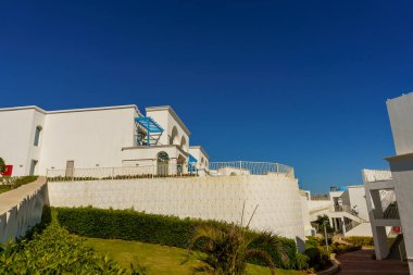 Mavi çatılı ve mavi balkonlu beyaz bir bina. Bina yemyeşil bir çimenlikle çevrili.