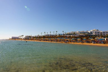 Bir sürü şemsiyeli ve insanlı bir sahil. Su berrak ve gökyüzü mavi.