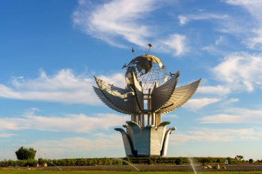 Üstünde kanatlı bir kuş olan büyük bir küre heykeli. Kuş bir çeşmeyle çevrili ve gökyüzü açık ve mavi