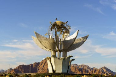 Kanatları ve kuşları olan büyük, süslü bir küre heykeli. Gökyüzü açık ve mavi, ve arkaplandaki dağlar sahneye ihtişam ve ihtişam katıyor.