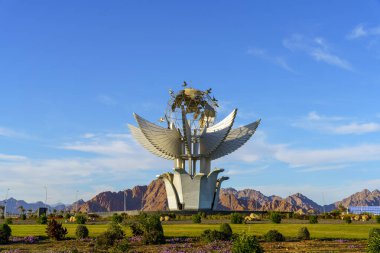 Açık mavi gökyüzü olan bir parkta büyük bir kuş heykeli var. Kuş, çalılar ve çiçekler de dahil olmak üzere bir sürü yeşillikle çevrilidir. Huzurlu ve huzurlu bir sahne.