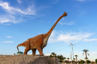 Yeşil bir binanın önünde bir dinozor heykeli duruyor. Gökyüzü mavi ve arka planda palmiye ağaçları var