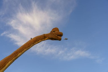 Ağzı açık bir dinozor ve bir kuş uçuyor. Gökyüzü mavi ve arka planda bazı bulutlar var