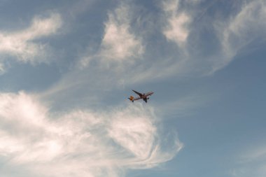 Bulutlu bir gökyüzünde bir uçak uçuyor. Gökyüzü çoğunlukla mavi ve bazı bulutlar etrafa saçılmış. Uçak alçaktan uçuyor, sanki bulutların arasında uçuyor gibi.