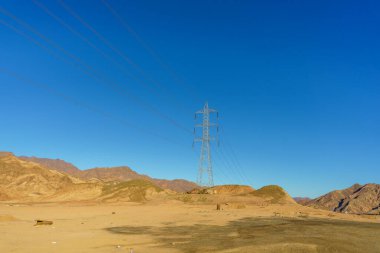 Bir güç hattı kulesi çölün ortasında. Gökyüzü açık ve mavi. Kule uzun ve içinden bir sürü kablo çıkıyor.