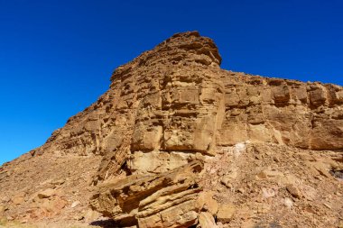 Arka planda mavi gökyüzü olan kayalık bir yamaç. Kayalar pürüzlü ve pürüzlü, sahneyi sert ve vahşi gösteriyor. Mavi gökyüzü arasındaki zıtlık