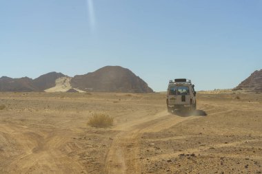 Çölde toprak bir yolda bir araba sürüyor. Gökyüzü açık ve güneş parlıyor.