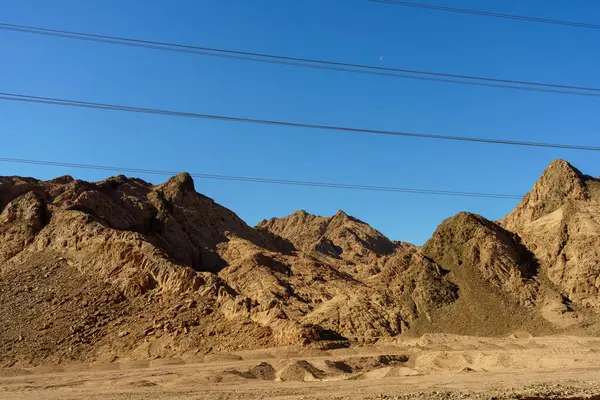 Arka planda açık mavi gökyüzü olan bir dağ sırası. Gökyüzü elektrik hatlarıyla dolu.