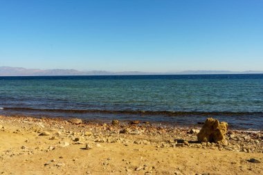 Arka planda büyük bir su kütlesi olan kayalık bir sahil. Su sakin ve gökyüzü berrak.