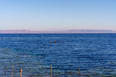 Okyanus sakin ve mavidir, suda birkaç kişi vardır. Gökyüzü berrak ve parlak. Huzurlu ve huzurlu bir atmosfer yaratıyor.
