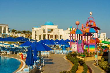 Mavi şemsiyeli ve pembe slaytlı renkli bir oyun alanı. Oyun alanı sandalyeler ve şemsiyelerle çevrili ve bölgede birkaç kişi var.