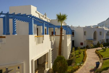 Önünde mavi tenteli ve palmiye ağaçları olan bir sıra beyaz bina. Binalar yüksek ve balkonları var. Binaların önündeki patika çalı ve ağaçlarla kaplı.