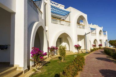 Önlerinde mavi tenteleri ve pembe çiçekleri olan bir sıra beyaz bina. Binalar yüksek ve camları kemerli.