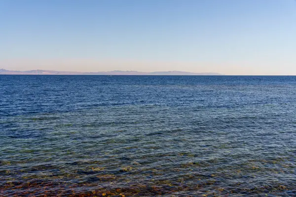 Okyanus sakin ve berrak, kıyıda birkaç küçük dalga kırılıyor. Gökyüzü parlak mavi, görünürde bulutlar yok. Huzurlu ve huzurlu bir sahne.