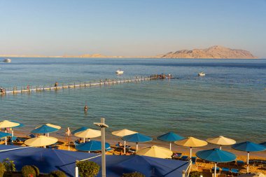 Bir sürü şemsiyeli ve insanlı bir sahil. Gökyüzü mavi ve su sakin.