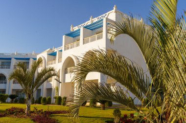 Önünde mavi aksanlı ve palmiye ağaçları olan büyük beyaz bir bina. Binanın mavi bir tentesi ve balkonu var.