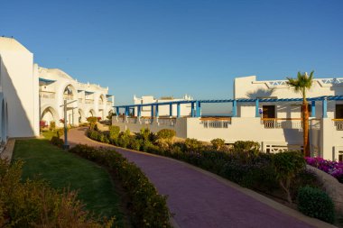 Mavi çatılı bir binaya çıkan patikalı bir yürüyüş yolu. Geçit ağaçlar ve çalılarla kaplı.