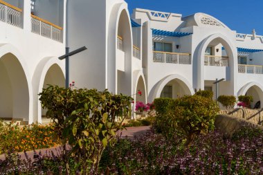 Mavi kemerli beyaz binalar, Akdeniz tarzı mimari izlenimi veriyor. Bu binaların önünde küçük yeşil bir çalı var.