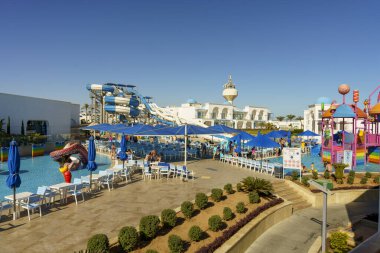 Üzerinde mavi bir gökyüzü olan büyük bir açık su parkı. Misafirlerin dinlenmesi ve su parkının keyfini çıkarmaları için kurulmuş birkaç şemsiye ve sandalye var.