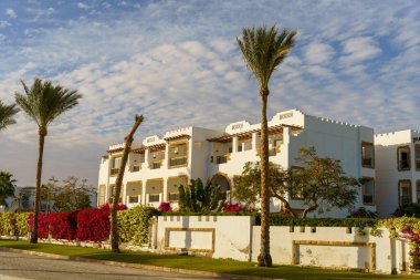 Önünde beyaz çitleri ve palmiye ağaçları olan beyaz bir bina. Binanın bir sürü penceresi var ve yemyeşil çimlerle çevrili. Gökyüzü bulutlu.