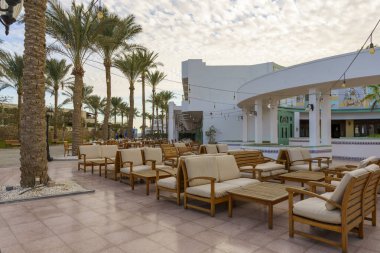 Bir sürü ahşap sandalye ve kanepesi olan büyük bir avlu. Sandalyeler sıra sıra dizilir ve kanepeler çeşitli yerlere serilir. Avlu palmiye ağaçları ile çevrilidir. Sahne davetkar.