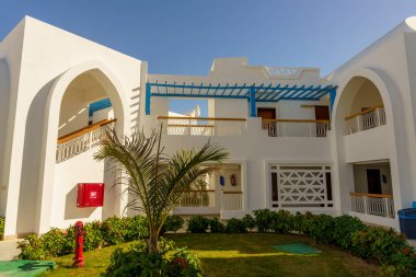 Önünde kırmızı kutu ve mavi aksanlı beyaz bir bina. Binada palmiye ağacı ve yangın musluğu olan bir avlu var.