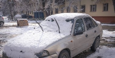Bir araba karla kaplı ve silecekleri açık. Araba bir otoparka park edilmiş.