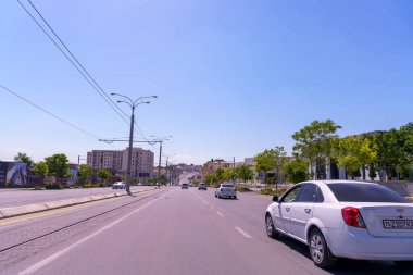 Beyaz bir araba caddeden geçiyor. Yolda başka birkaç araba daha var. Gökyüzü açık ve mavi