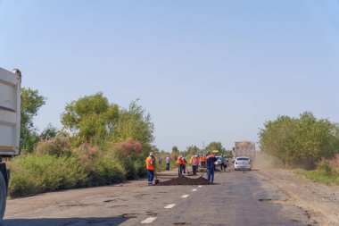 Bir grup işçi bir yolu onarıyor. Arka planda birkaç kamyon var. Kamyonlardan biri çöp konteynırı taşıyor.