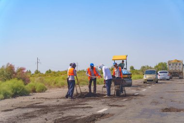Bir grup adam bir yol üzerinde çalışıyor. Adamlardan biri sarı bir şapka takıyor.