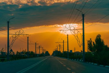 Arka planda günbatımı olan bir yol. Güneş batıyor ve gökyüzü turuncu. Arka planda bir sürü elektrik hattı var.