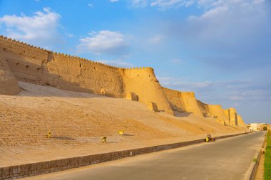 Yan tarafında duvar olan bir yol. Duvar taştan yapılmış. Yolda yürüyen iki kişi var.