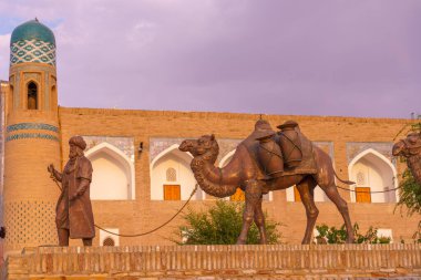 Bir deve heykeli bir binaya zincirlenir. Heykelin yanında bir adam duruyor. Heykel tuğla bir duvarda.