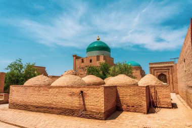 Tepesinde kubbe olan bir bina ve tuğladan yapılmış bir duvar. Binanın önünde ağaçlar var.
