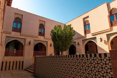 Ortasında ağaç olan bir avlu. Kemerli pencereli birkaç bina var. Avlu tuğla bir duvarla çevrili.