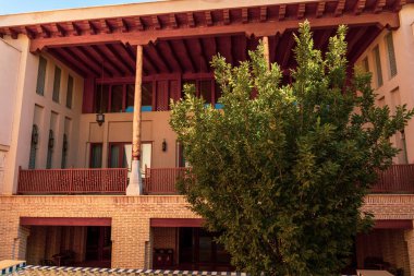 Binanın önünde bir ağaç büyüyor. Ağaç yeşil ve yaprakları var. Bina kırmızı ve balkonu var.