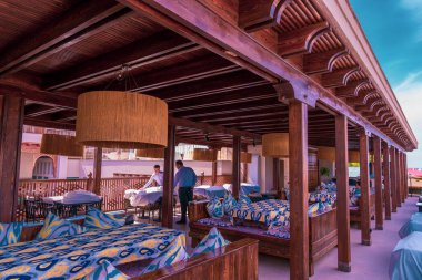 Bir sürü masası ve sandalyesi olan bir restoran. Restoranda bir adam yürüyor. Sandalyesi olan bir sürü masa var ve bir sürü insan oturuyor.