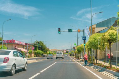 Bir adam bir binanın önündeki kaldırımda ayakta duruyor. Sokakta birkaç araba var, biri beyaz diğeri siyah araba. Sokaklar ağaçlarla dolu.