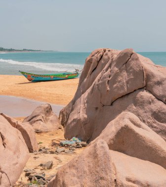 Sahilde büyük bir kayanın yanında bir tekne oturuyor. Kaya, etrafında birkaç küçük kaya bulunan büyük bir kayadır. Kumsal kumlu ve su sakin. Huzurlu ve huzurlu bir sahne.