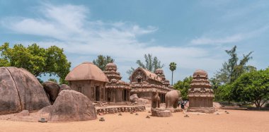 Arkasında büyük bir kaya olan bir grup bina. Bölgede dolaşan insanlar var. mahabalipuram