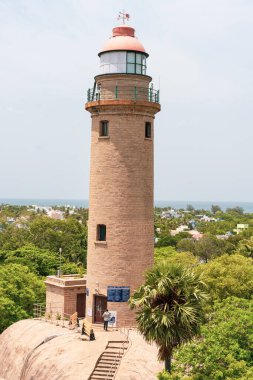 Okyanusa bakan bir tepede uzun bir deniz feneri var. Deniz feneri ağaçlar ve bir palmiye ağacıyla çevrilidir. Deniz fenerinin tabanında yürüyen insanlar var.
