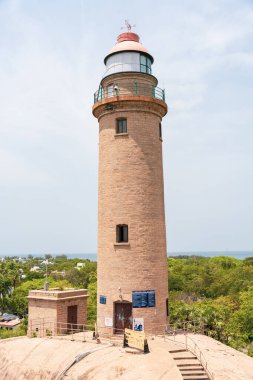 Üzerinde mavi bir işaret olan uzun bir deniz feneri. Deniz feneri bir tepenin üzerinde ve okyanus manzarası var.