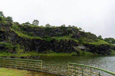 Yeşil ve sarı bir çit bir su kütlesini sarar. Su sakin ve temiz. Çevresi yemyeşil ve yemyeşil. Gökyüzü bulutlu, ortama huzurlu ve huzurlu bir atmosfer veriyor.