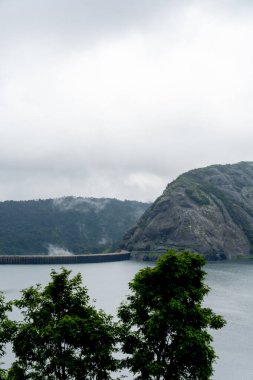 Önünde bir su kütlesi olan bir dağ sırası uzaklıktan görülebilir. Gökyüzü bulutlu, karamsar bir atmosfer yaratıyor