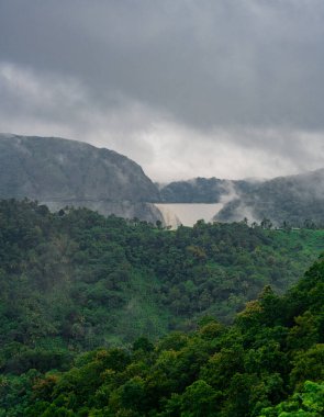 Önünde şelale olan bir dağ sırası. Gökyüzü bulutlu ve ağaçlar yemyeşil